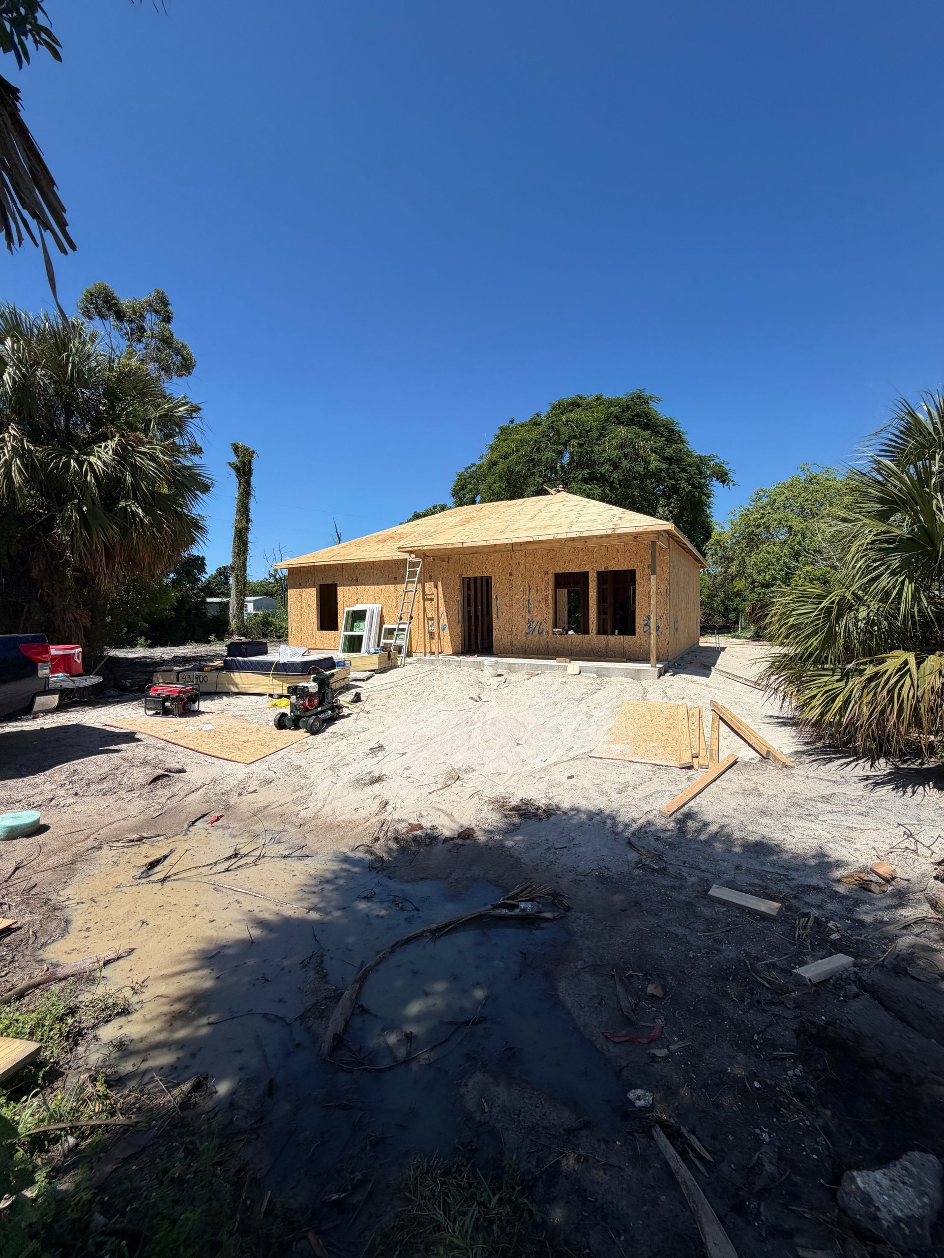 New house under construction with cork or natural material exterior, surrounded by palm trees and sandy ground on a sunny day