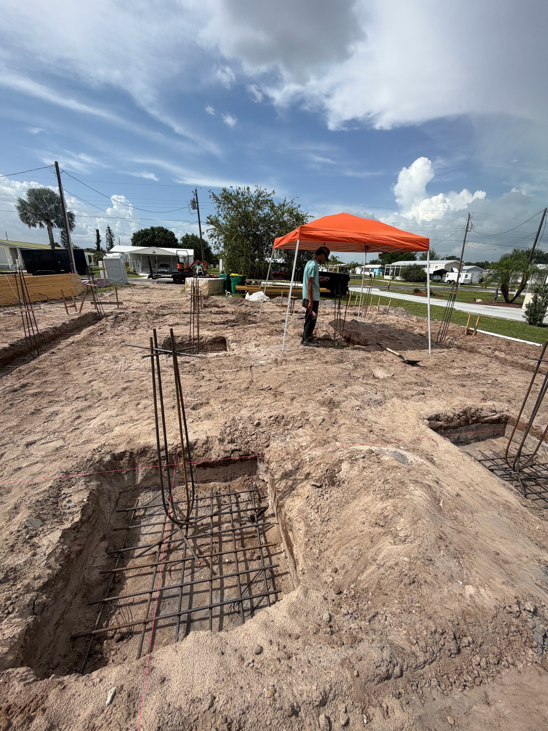 Construction site with foundation holes and steel reinforcement bars under orange canopy tent with blue sky
