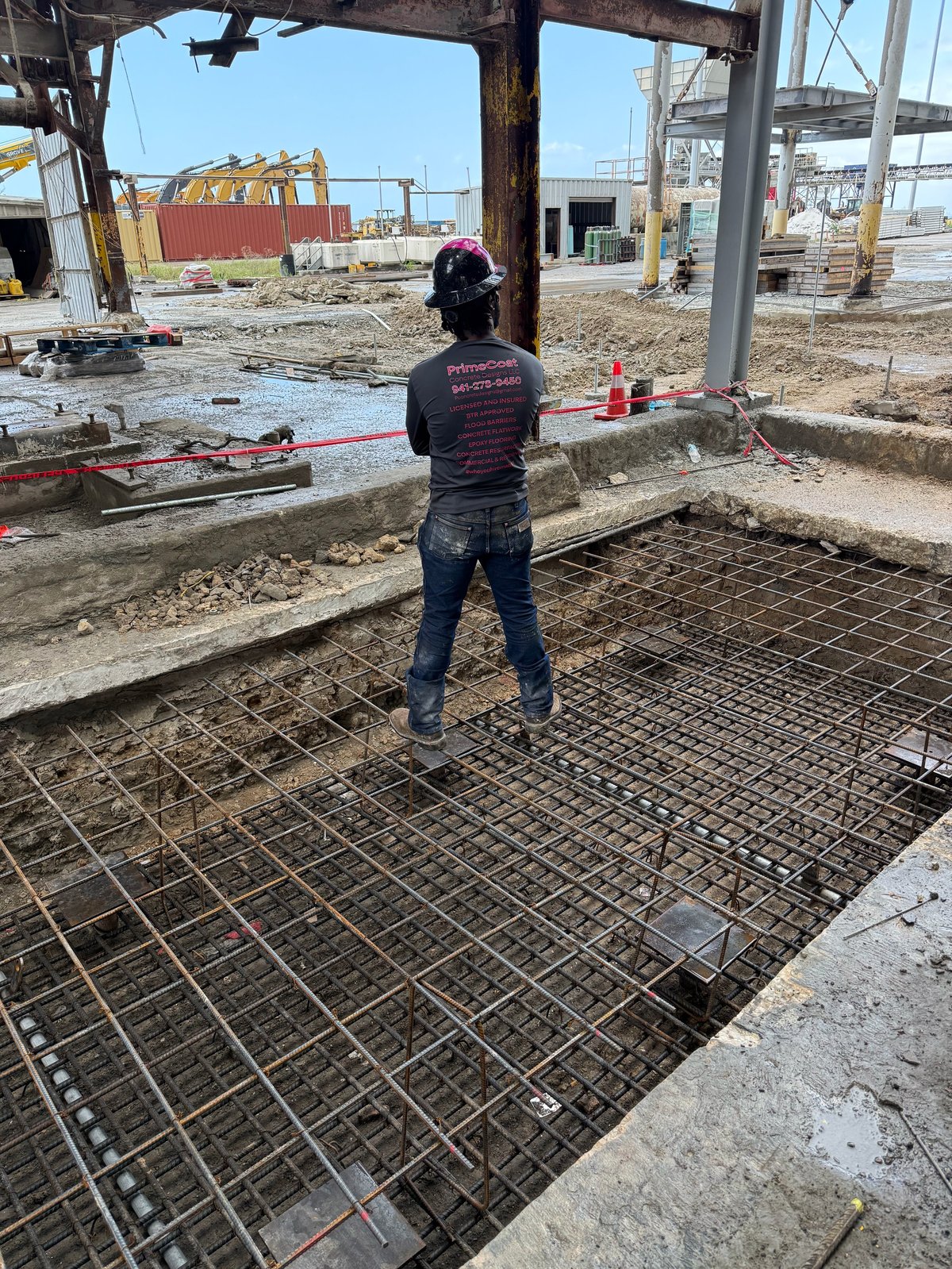 Construction worker standing on rebar mesh at an active building site with heavy equipment and industrial structures in the background
