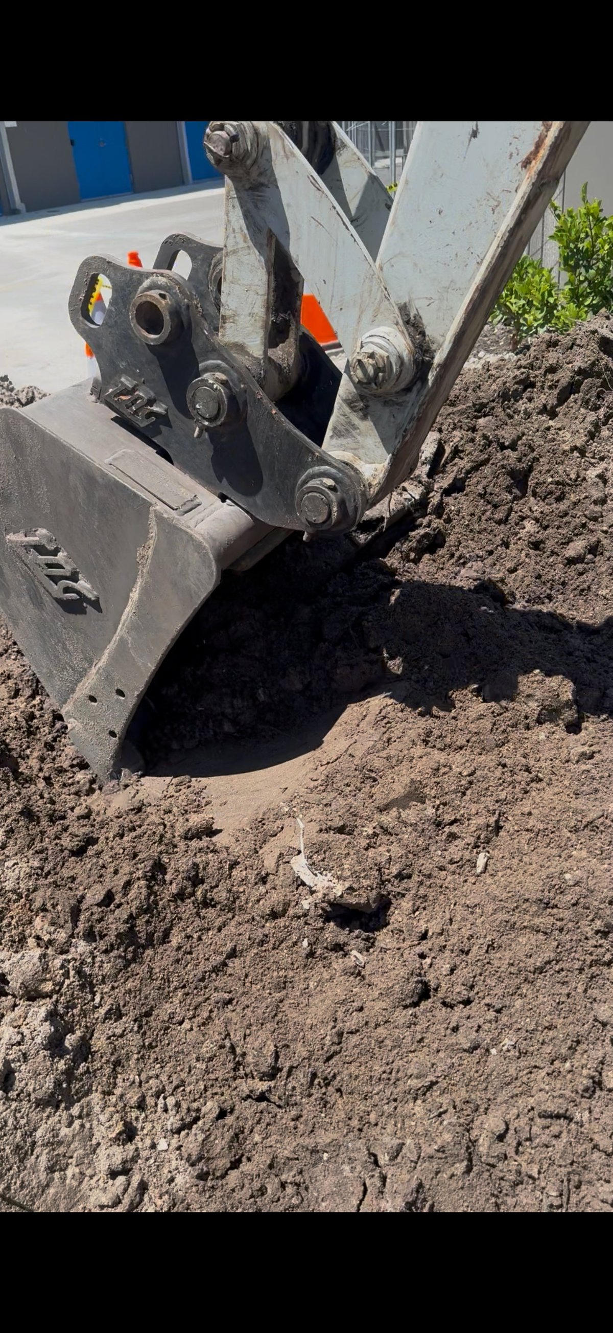 Excavator bucket digging into brown soil at a construction site with orange safety markers visible