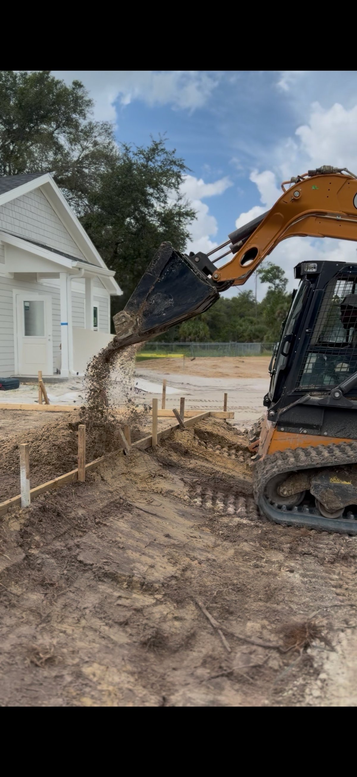 Orange excavator dumping soil near a white house under construction on a sunny day