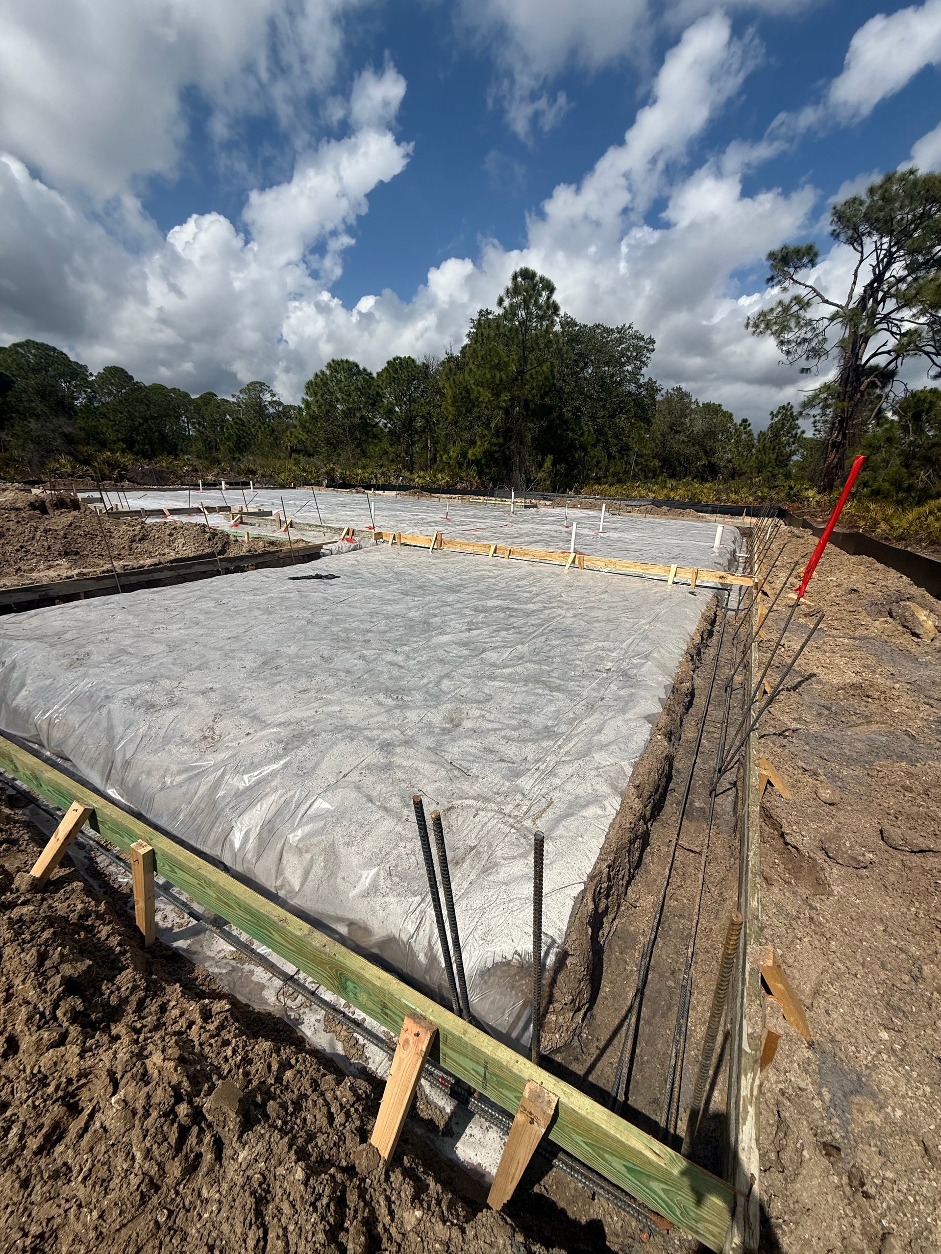 Construction site with concrete foundation, plastic sheeting, wooden forms, and rebar reinforcement under blue sky with trees