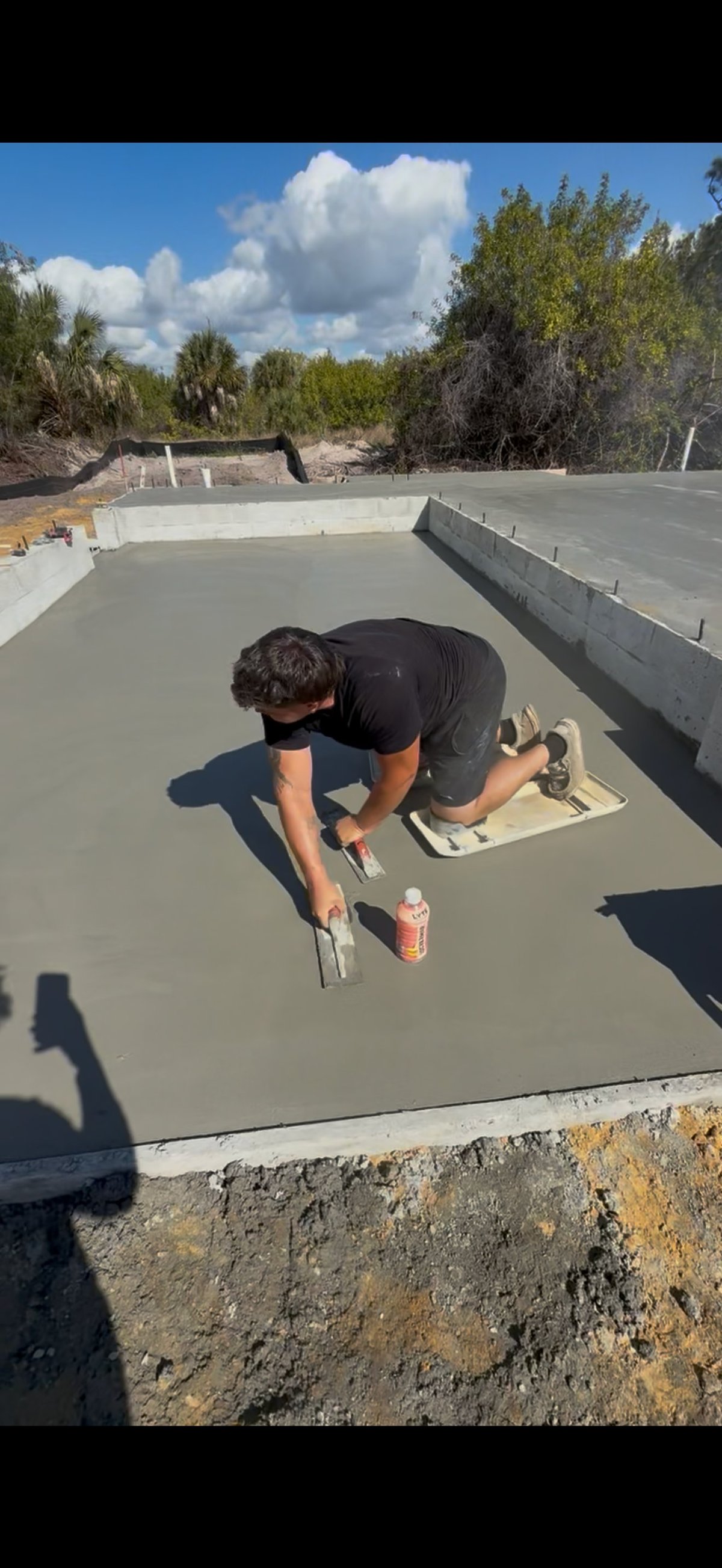 Worker applying concrete finish to pool deck under bright Florida sunshine with palm trees and clouds visible in background