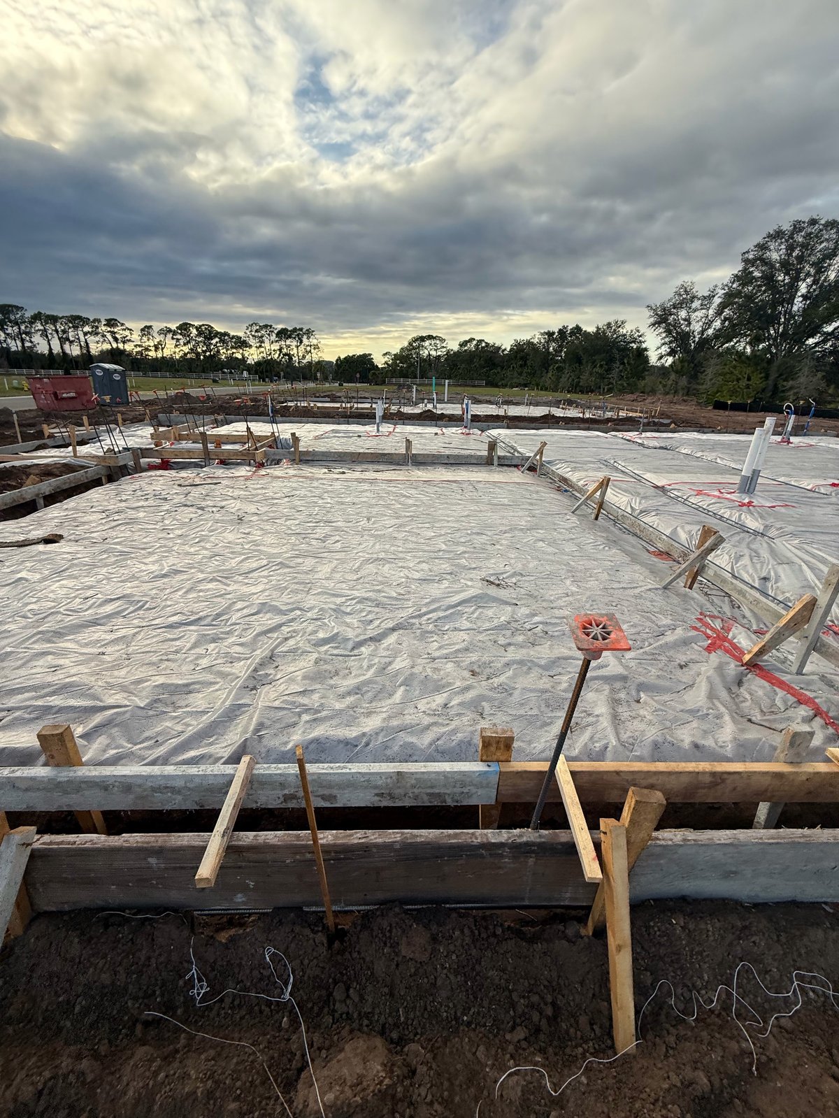 Construction site with concrete foundation areas under cloudy sky, showing wooden formwork and safety markers