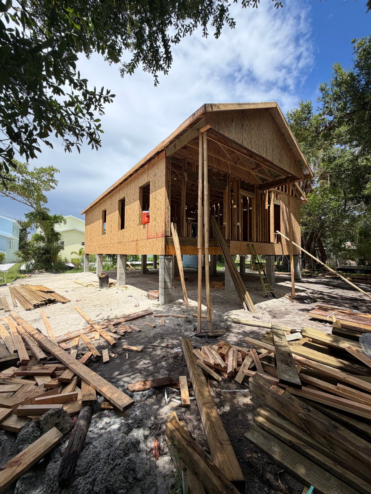 Brick house under construction elevated on wooden pilings with framing exposed, surrounded by lumber and materials on sandy ground
