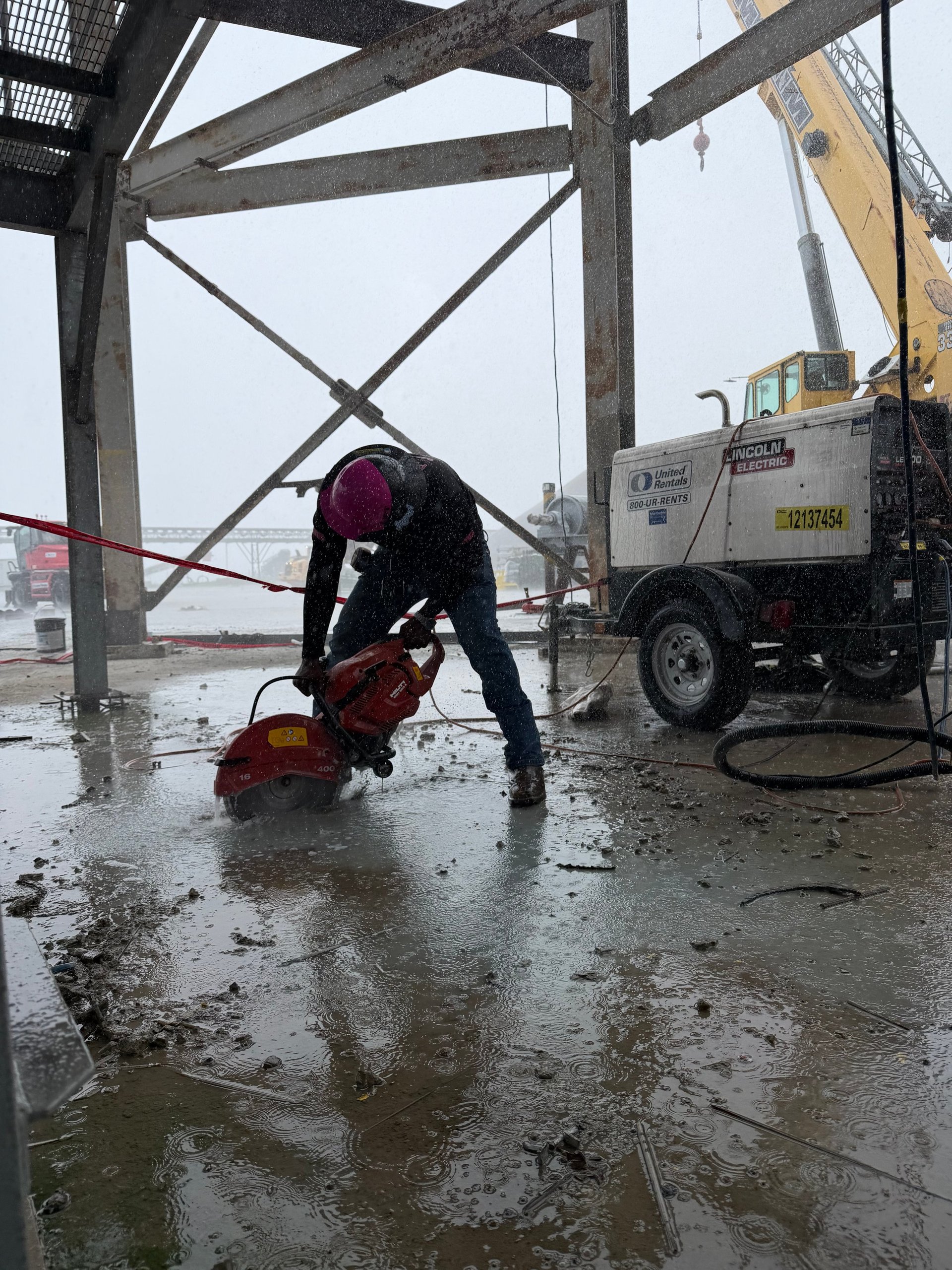 Worker operating jackhammer on wet concrete under metal bridge structure with cement truck nearby
